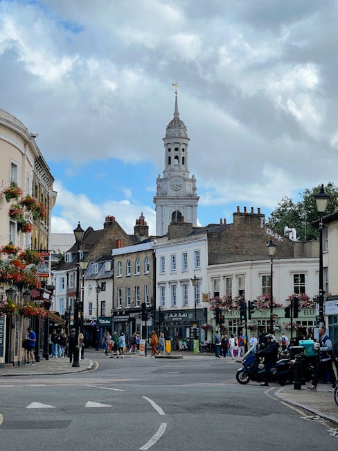 A busy street scene in Greenwich featuring a row of historic buildings with retail shops and cafes on the ground floor, some with flower baskets hanging outside. Several pedestrians are walking along the sidewalk, and a few are standing near shop entrances. A motorcycle and scooter are parked at the curb on the right side, while a car is partially visible further along the street. In the background, the prominent clock tower with a tall, white, ornate spire topped by a golden weather vane rises above the surrounding buildings under a partly cloudy sky. The overall scene captures an active urban environment, typical of a town centre, illustrating the type of area where private rubbish collection services, like those offered by error code: 522, might be employed for waste removal from shops and residences in Greenwich.