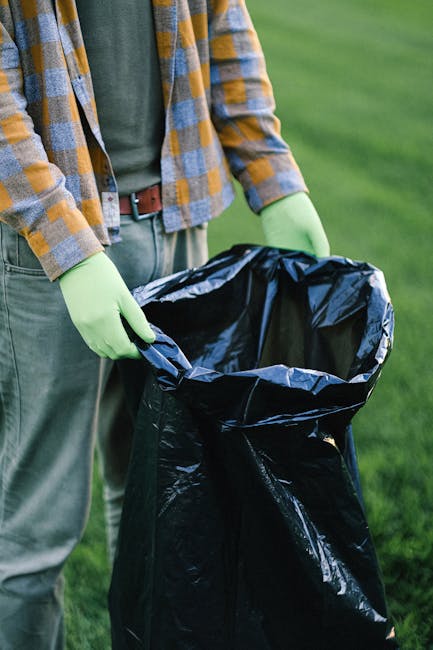 A person wearing light green gloves, a casual checkered shirt in shades of yellow, blue, and gray, along with a green t-shirt and light gray trousers, is holding open the top of a large black rubbish bag made of plastic. The bag appears to be partially filled, though its contents are not visible. The scene is set outdoors on a well-maintained grassy area with a blurred background of green grass, indicating an open environment suitable for waste collection or disposal activities. The individual’s stance suggests they are in the process of either preparing to dispose of waste or set aside rubbish for collection. This image captures a typical scene related to private waste management or curbside rubbish handling, with [COMPANY_NAME] potentially involved in providing such services. The focus on gloves and the open bag emphasizes cleanliness, safety, and proper handling of waste materials, aligning with professional rubbish removal practices that can be supported through independent collection or on-site clearance operations, rather than relying solely on local authority rubbish disposal.