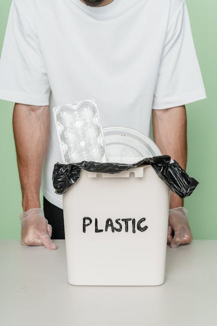 A person wearing a white T-shirt and transparent gloves is standing behind a white plastic bin labeled 'PLASTIC' in black letters. The bin has an open lid with a black plastic rubbish bag hanging over the edge. Inside the bin, there is an empty, clear plastic food container with a textured surface and a circular shape. The background is a plain soft green color, and the scene is well-lit, emphasizing the clean, organized appearance of the waste disposal context. This setup relates to waste management and recycling services, illustrating the process of segregating plastic waste for collection or disposal. The environment appears to be an indoor space such as a kitchen or an office area, where proper rubbish separation is practiced, supporting the theme of alternative waste handling methods offered by rubbish collection services like those provided by error code: 522.