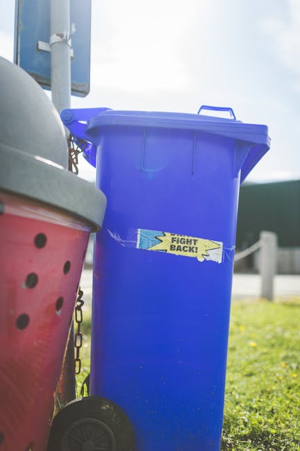 A close-up view of a large green waste bin on wheels, placed on a paved surface next to a dark brick wall. The bin is partially filled with red and black bags of garden or general waste, labeled with markings indicating they contain flexible garden hoses or watering accessories. The bin appears to be used for private rubbish collection and is positioned next to another similar bin. There is a white plastic bin liner inside with visible text and a partially open cardboard box resting against the side of the bin, with a small section of a metal ramp or grate on the ground nearby. The scene suggests an outdoor setting, possibly a driveway or courtyard, used for interim waste storage before collection, reflecting an independent disposal or rubbish removal service in Greenwich.