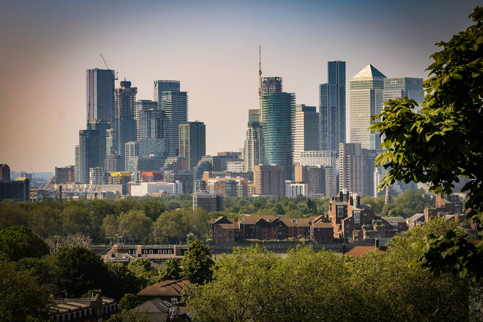 A city skyline featuring numerous modern high-rise buildings constructed from glass and steel, with varying heights and architectural styles. Some buildings are under construction, evidenced by visible cranes, and the tallest structures include office towers and a prominent pylon. In the foreground, there is a quiet suburban area with low-rise brick houses surrounded by lush green trees, creating a natural contrast to the dense urban landscape behind. The scene appears to be taken during daylight hours with clear, slightly hazy weather, casting soft shadows and illuminating the buildings with natural light. The upper right corner partially includes the leafy branches of a large tree, framing the view. This scene illustrates the juxtaposition of private residential spaces with the expansive city centre, aligning with themes of alternative waste handling and independent collection methods in urban environments, as highlighted in the context of rubbish removal services such as those offered by error code: 522.