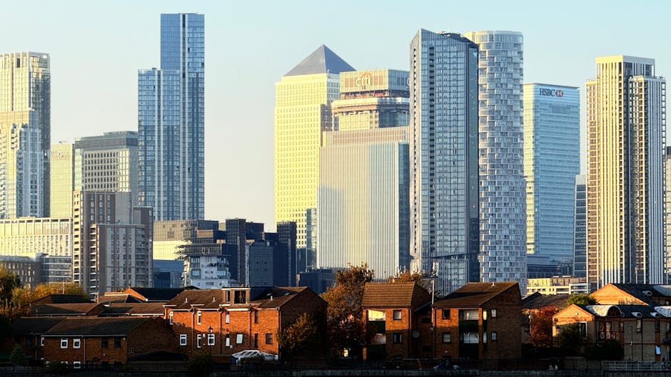 A cityscape featuring a mix of tall modern skyscrapers with glass facades and glass panels reflecting sunlight, situated behind a row of small, red-brick residential houses with tiled roofs. The skyscrapers vary in height and design, with some displaying corporate logos such as HSBC and HSBC UK, and include office buildings with reflective windows. The foreground shows the houses with windows, some with small balconies, and trees visible around them, creating a contrast between urban residential and commercial architecture. The scene is captured during daylight with clear skies, highlighting the differences in architectural materials and styles, and reflecting the potential for alternative waste handling solutions like independent rubbish collection services in densely built environments.