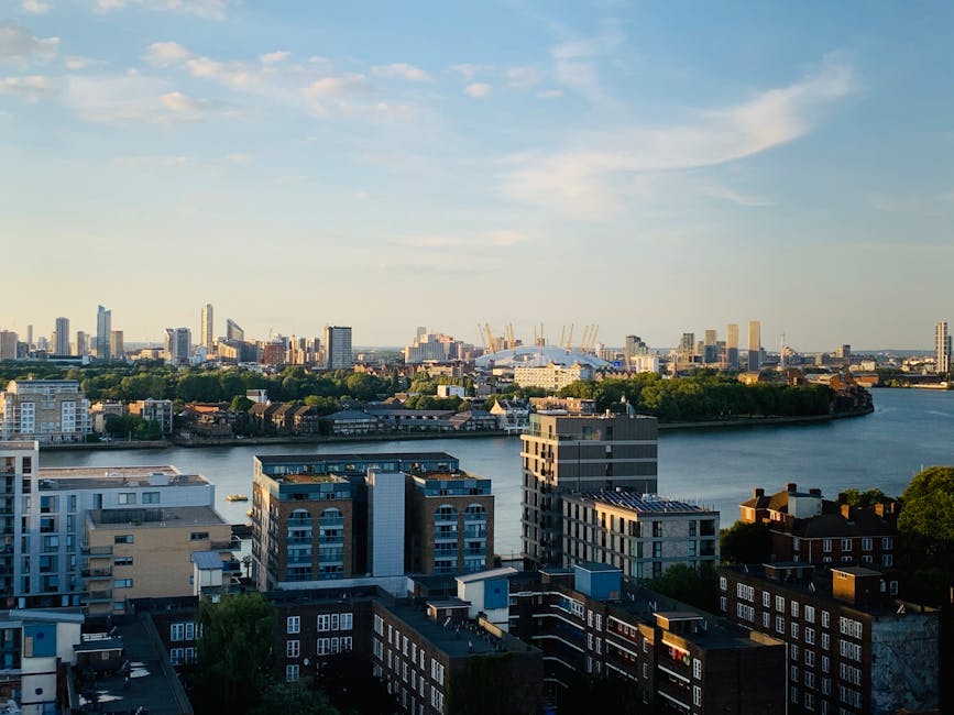 A city skyline featuring numerous modern high-rise buildings constructed from glass and steel, with varying heights and architectural styles. Some buildings are under construction, evidenced by visible cranes, and the tallest structures include office towers and a prominent pylon. In the foreground, there is a quiet suburban area with low-rise brick houses surrounded by lush green trees, creating a natural contrast to the dense urban landscape behind. The scene appears to be taken during daylight hours with clear, slightly hazy weather, casting soft shadows and illuminating the buildings with natural light. The upper right corner partially includes the leafy branches of a large tree, framing the view. This scene illustrates the juxtaposition of private residential spaces with the expansive city centre, aligning with themes of alternative waste handling and independent collection methods in urban environments, as highlighted in the context of rubbish removal services such as those offered by error code: 522.