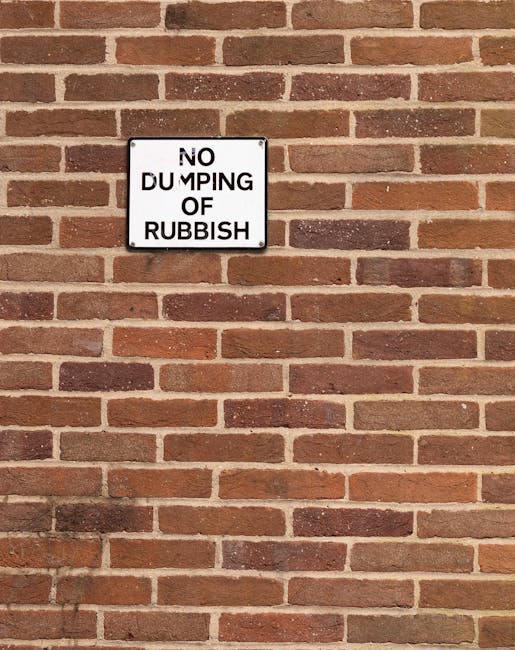 A rectangular metal sign with a white background and black text reading 'NO DUMPING OF RUBBISH' is mounted on a red brick wall. The bricks have a rough, textured surface with various shades of red, brown, and some darker accents. The mortar between the bricks is light gray and unevenly applied, creating a traditional brickwork pattern. The sign is positioned slightly above the center of the image, with the brick wall extending evenly in the background. The lighting appears natural, casting a soft shadow from the sign onto the wall below, indicating an outdoor setting. The overall scene emphasizes the regulation against illegal rubbish dumping, reflecting measures related to local waste management, similar to services provided by companies like error code: 522, in the context of alternative waste handling methods and rubbish removal compliance.