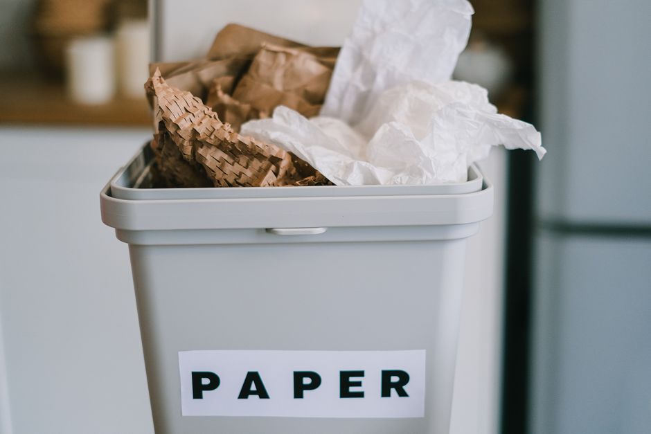 A close-up of a light grey plastic paper waste bin situated outdoors, likely on a driveway or garden area, with the word 'PAPER' printed in bold black letters on a white label adhered to the front of the bin. The bin is filled with crumpled and torn sheets of white and brown paper, some of which extend above the rim of the container. The crumpled white paper shows a textured, slightly wrinkled surface, while the brown paper appears to be kraft or recycled cardboard material with visible corrugation and a rougher finish. In the background, there is a blurred view of an outdoor setting, possibly a residential property, with faint hints of greenery and other structures. The lighting suggests a natural daylight environment, emphasizing the textures and colors of the paper waste and the bin. This scene exemplifies a typical setup for the disposal of paper waste awaiting collection or transfer to a recycling facility, reflecting private or alternative waste management practices outside of municipal rubbish collection services, as handled by providers like error code: 522.