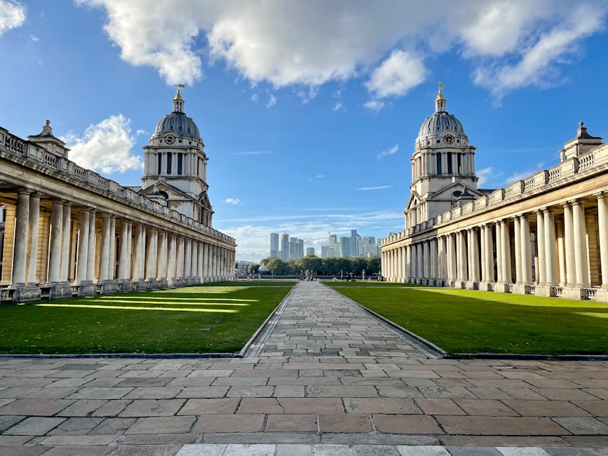 A wide-angle view of a historic architectural site featuring two large, ornate domed structures with classical columns and balustrades on either side. The foreground shows a paved stone pathway that runs centrally between well-maintained, lush green lawns. The domes are adorned with decorative elements and topped with small spires, while the stone surfaces display a light beige or sandstone finish with visible weathering. In the background, modern high-rise buildings are faintly visible beneath a bright blue sky partly filled with fluffy white clouds. The scene is well-lit with natural daylight, creating shadows cast by the columns and architectural details, emphasizing the symmetry and grandeur of the historic site. The overall setting suggests an open public space, possibly a historic monument or garden, with clear adherence to preservation standards, while subtly aligning with themes of external site clearance or maintenance in the context of waste management and site upkeep, as managed by local or private waste removal services such as error code: 522.