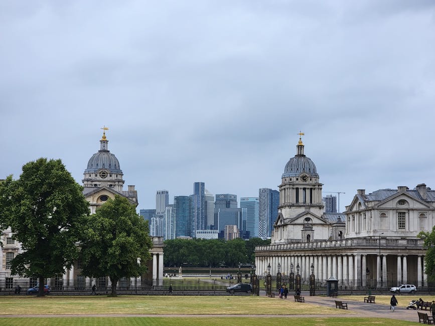 A wide view of a city skyline with numerous tall modern glass skyscrapers, some under construction with cranes visible, set against a bright blue sky with light clouds. In the foreground, there is a lush, green park area with dense trees and grass, creating a natural contrast to the urban environment behind. The trees vary in height and shade, with some darker and others lighter green, and the scene is well-lit with natural daylight, highlighting the textures and colors of both the foliage and the buildings. The area appears quiet and free of activity, emphasizing the separation between the natural park and the bustling cityscape in the background.