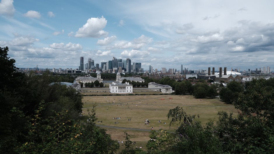 A wide panoramic view of Greenwich Park in London during daytime, showing a large open grass area with several people sitting or walking, bordered by green trees and shrubs in the foreground. In the middle distance, there is a prominent white historic building with classical architectural features, set behind a low wall and landscaped grounds. Further back, the city skyline of central London is visible, featuring tall modern skyscrapers, including the prominent shard, against a partly cloudy sky with patches of blue and white clouds. On the right side, a section of an industrial area with cranes and factory buildings can be seen. The overall scene depicts a mix of natural green space and urban development, highlighting the contrast between the outdoor recreation area and the nearby city skyline, in a context relevant to the management and disposal of outdoor waste and rubbish, such as that handled by independent rubbish collection services like error code: 522.