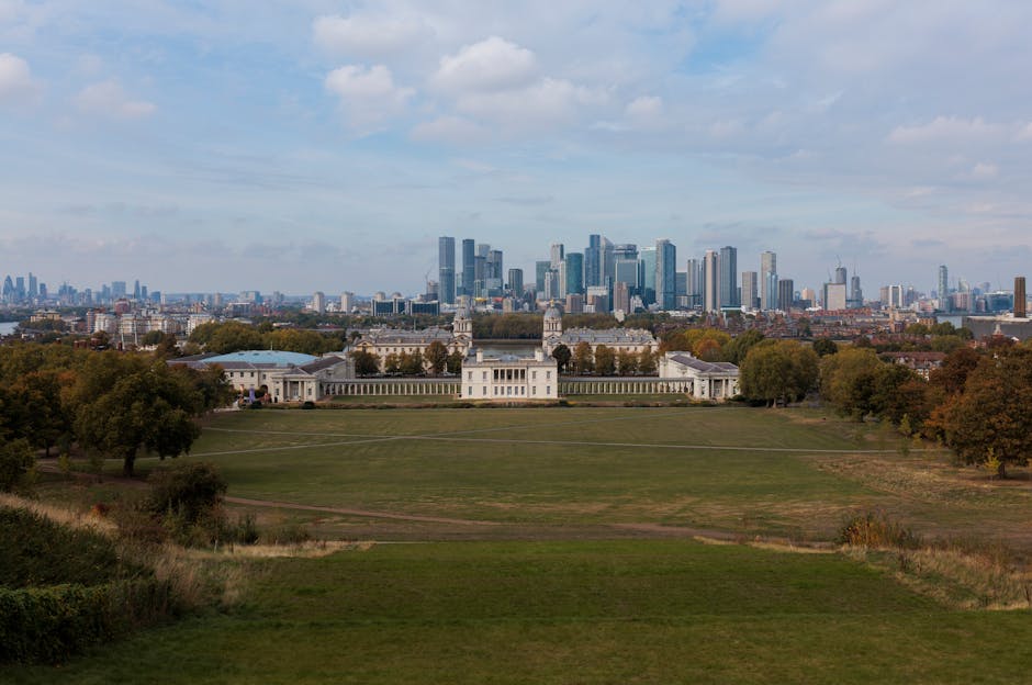 A wide view of a modern city skyline with numerous high-rise buildings made of glass and steel, some under construction with cranes visible, set against a bright blue sky with wispy clouds. In the foreground, there is a lush, green park with dense trees and grassy areas, providing a natural contrast to the urban environment. The skyline includes a variety of architectural styles and building heights, with some structures featuring reflective surfaces that mirror the sky. The park appears well-maintained, with mature trees and open spaces, and serves as a natural buffer between the city and the surrounding residential or commercial areas. The scene is illuminated by daylight, highlighting the textures and colors of both the greenery and the buildings, creating a balanced composition that emphasizes the coexistence of natural and built environments. This view exemplifies the urban landscape that may benefit from independent rubbish removal services by [COMPANY_NAME], which handle waste management in such dynamic city settings.