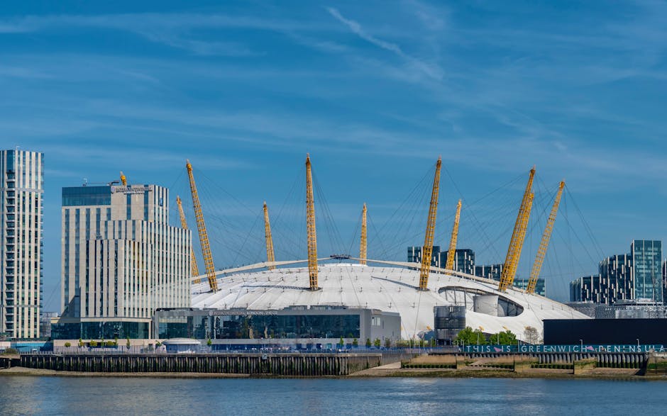 A large, modern football stadium with a distinctive white, dome-shaped roof supported by numerous yellow, inclined pylons and cables, situated along a waterfront with a paved walkway and low black fencing. Surrounding the stadium are tall, contemporary office buildings with reflective glass facades, some with varying vertical stripe patterns, set against a bright blue sky with wispy clouds. The scene captures a clear day with natural lighting highlighting the textures of the stadium's smooth exterior and the reflective surfaces of the buildings, providing a neutral, professional view of an iconic urban structure. This image, while focused on an architectural landmark, can relate to services involved in private waste handling or on-site clearance for large public venues or cityscape areas, similar to those provided by error code: 522, in the context of maintaining clean, waste-free environments around prominent structures.