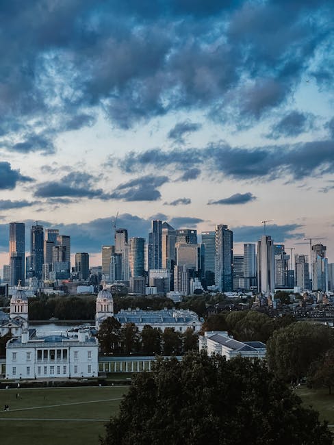 A panoramic view of a modern city skyline featuring numerous high-rise office buildings and residential towers made of glass and steel, reflecting the ambient daylight. The buildings vary in height, with some cranes visible atop the tallest structures, indicating ongoing construction. In the foreground, there is a historic white building with columns and a flat roof, situated within a park area that includes well-maintained lawns and grouped deciduous trees with dense, dark green foliage. The park's open grassy space extends towards the city, separated from the urban core by a layer of mature trees. The sky overhead is filled with dynamic cloud formations, featuring patches of blue sky peeking through, creating a contrast between the natural environment and the built environment below. This scene exemplifies a cityscape that might be associated with independent or private waste management, with the urban landscape seen from a vantage point that could align with local rubbish removal services covering residential and commercial areas.
