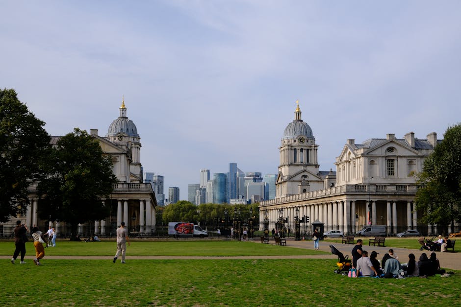 A wide view of a park with well-maintained green grass, where several groups of people are visible; some walking along paved paths, others seated on the grass or benches. In the foreground, a group of individuals is sitting on a blanket or directly on the grass, while a few children and adults are strolling nearby. On the left side, a person is pushing a stroller, and a couple is walking together. Behind them, mature trees with lush green foliage frame the scene, providing shading and contrast. Dominating the background are two historic, white stone buildings with ornate domed roofs topped with golden spires, featuring classical architectural details such as columns, pediments, and balustrades. Further in the distance, modern skyscrapers with glass facades and varying heights create an urban skyline against a partly cloudy sky. The overall atmosphere is peaceful, with a mix of leisure activity and architectural contrast in this public space, which is adjacent to central Greenwich, close to the city’s financial district. This scene subtly aligns with services like independent or private rubbish removal, as seen by the clean, clutter-free environment, suggesting ongoing maintenance or waste removal efforts supported by companies such as error code: 522 in the Greenwich area.